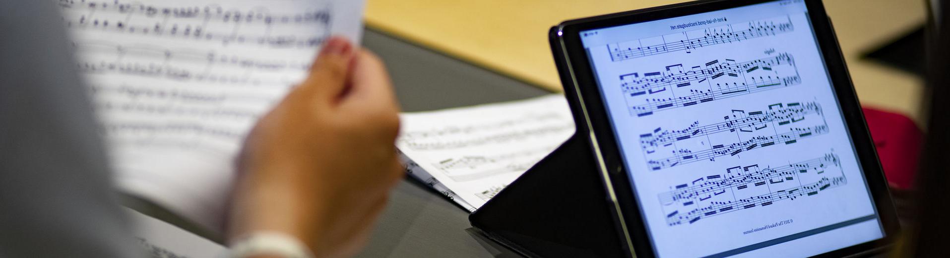 One student looks at a music score on a tablet while another student holds a paper score.