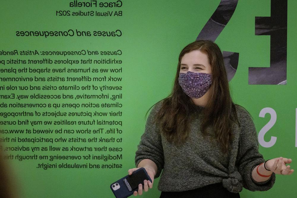 A masked Tyler student holds a phone as they stand in front of their work in a gallery.
